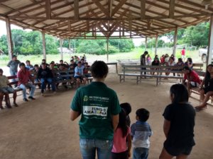 Homem de costas rodeado de crianças fala para grupo de pessoas em uma das terras indigenas