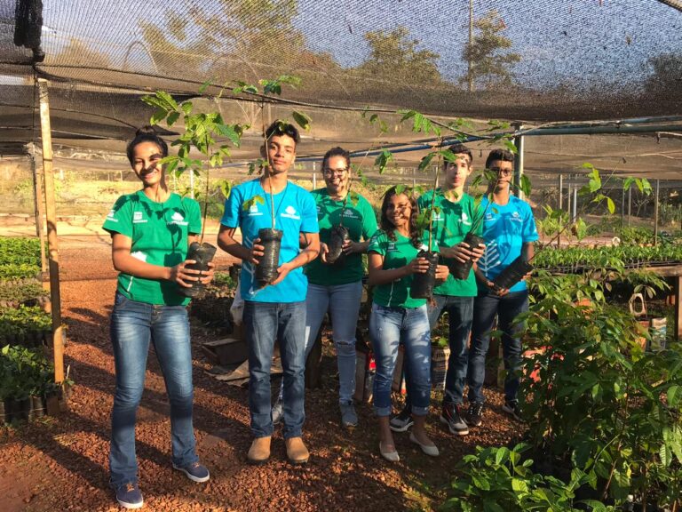 A foto mostra seis estudantes em um viveiro com mudas de árvores nas mãos.